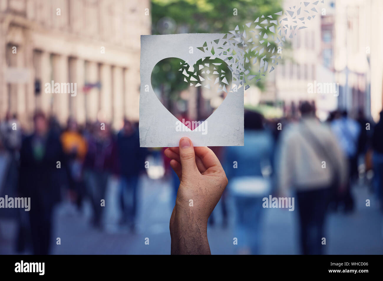 Health and wellbeing global issue as human hand holding a paper with heart symbol breaking into pieces over a crowded street background. Suffering myo Stock Photo
