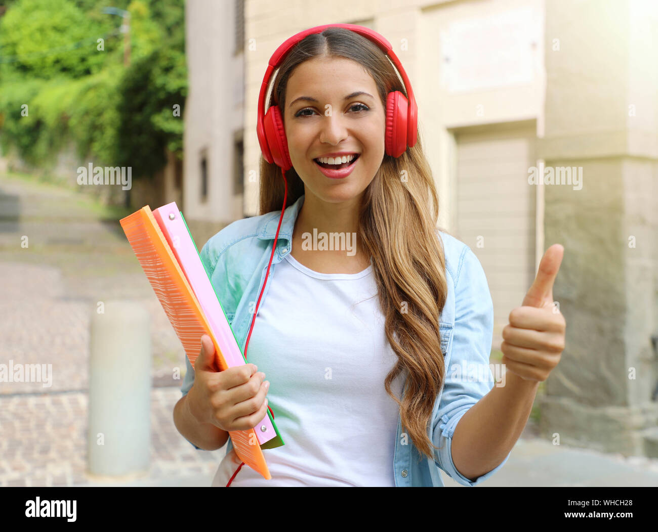 Happy student girl with headphones gives thumb up outdoors. Young woman doing student exchange programme. Stock Photo