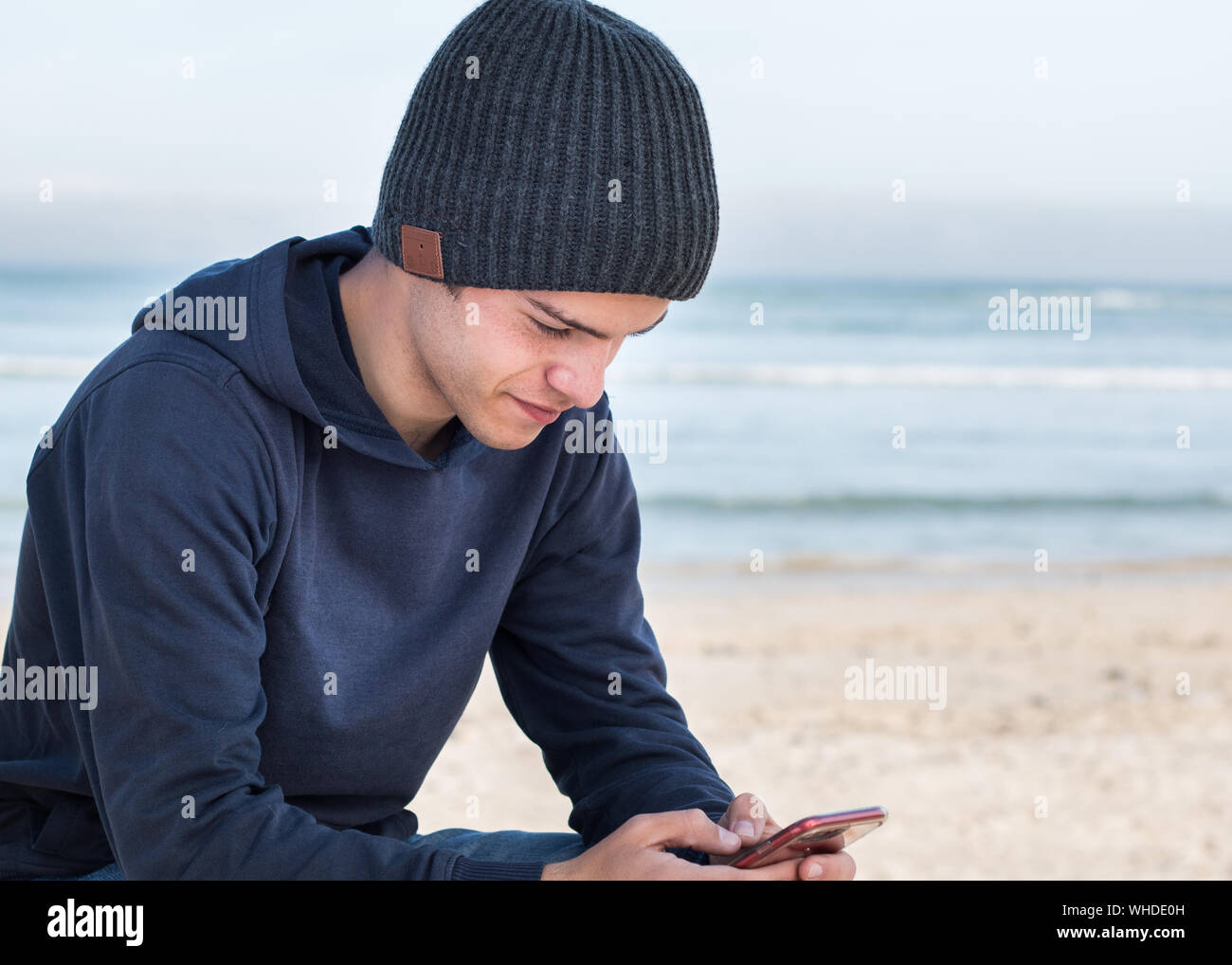 Male model wearing dark grey knitted beanie with Bluetooth speakers inside, listening to music with mobile smart phone outdoors. Looking at mobile dev Stock Photo