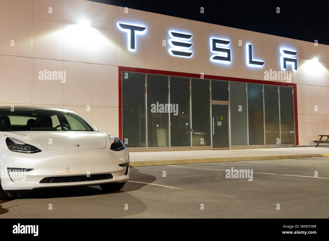 Tesla Model 3 parked in-front of an illuminated Tesla logo at a dealership at night. Stock Photo