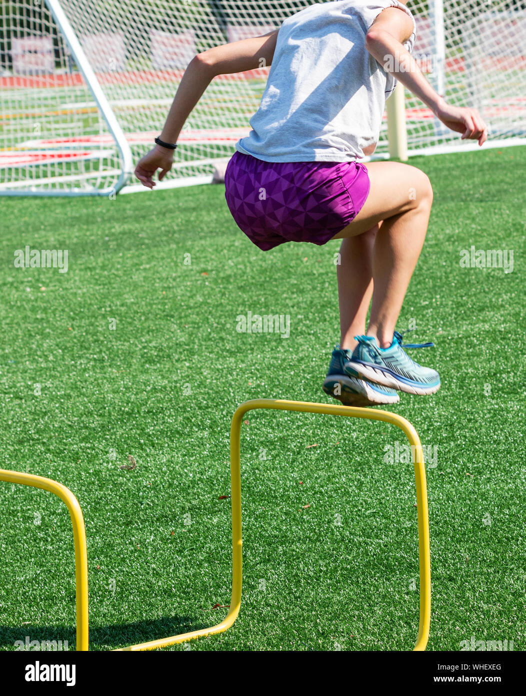 A high school runner wearing purple shorts is jumping over two foot yellow hurdles during strength and agility practice. Stock Photo