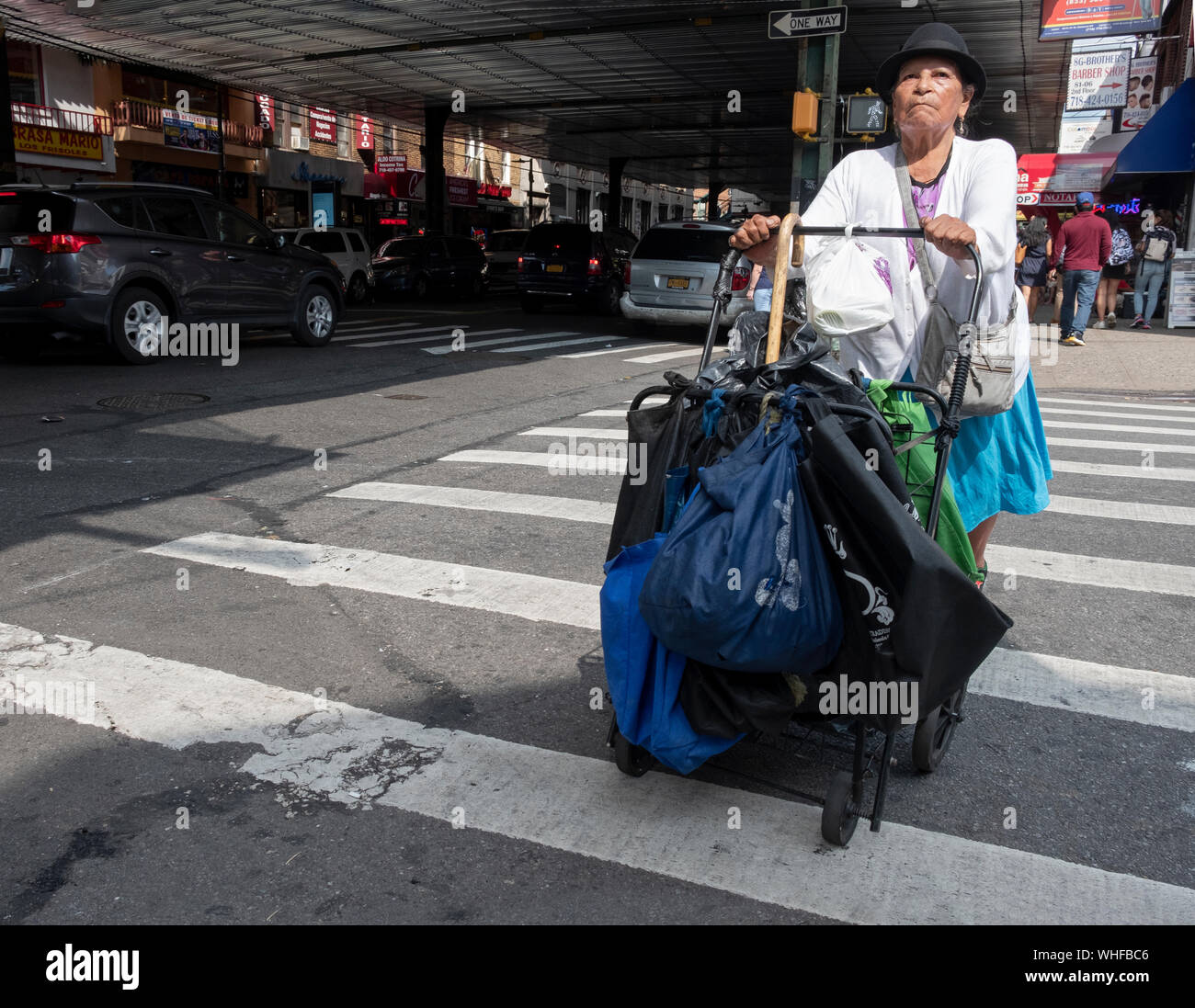 A woman in a hat, probaly Mexican, crosses the street pushing a cart. On Roosevelt avenue in Jackson Heights, Queens, New York City. Stock Photo