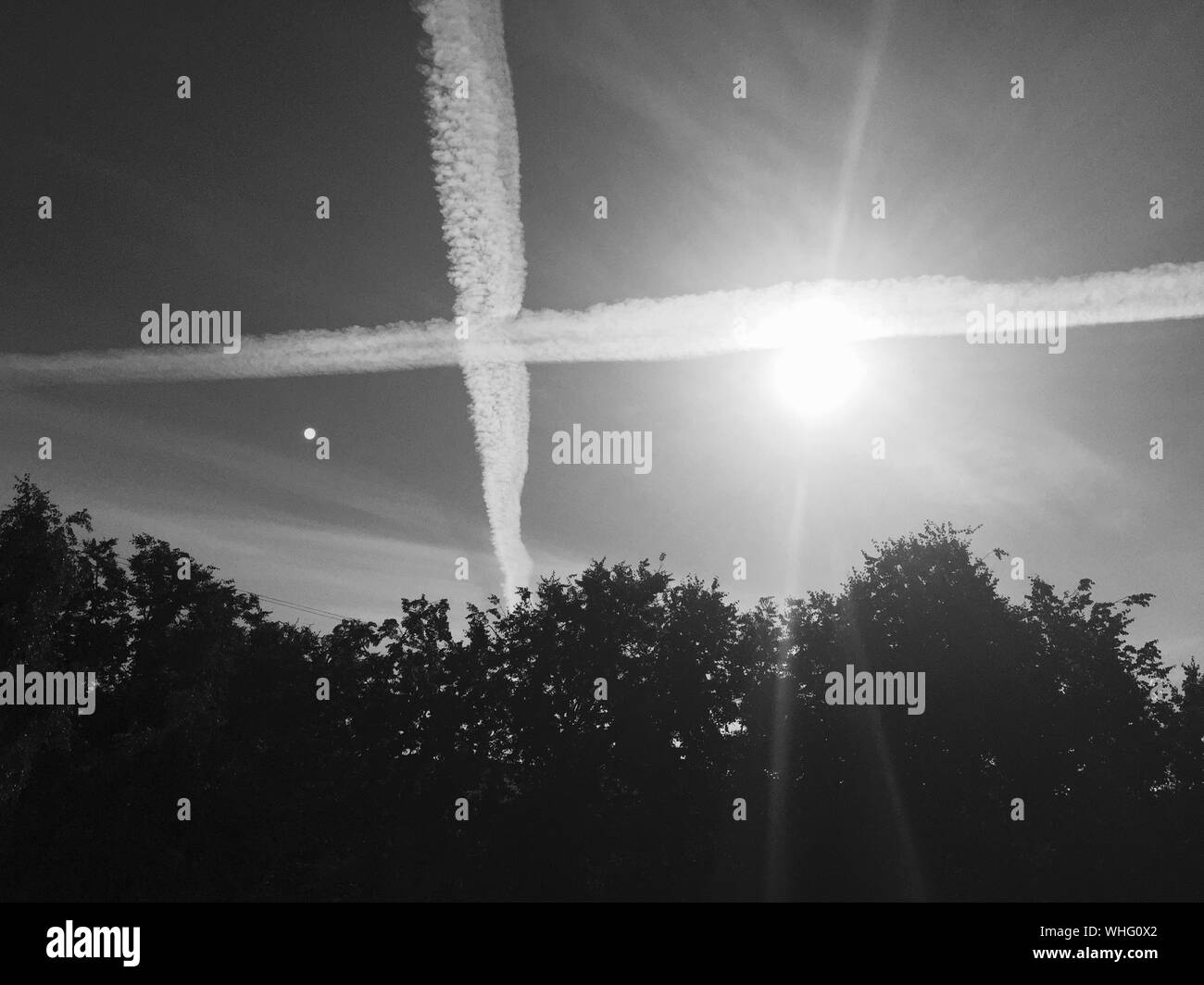 Low Angle View Of Vapor Trail And Sunlight Over Silhouette Trees Stock Photo
