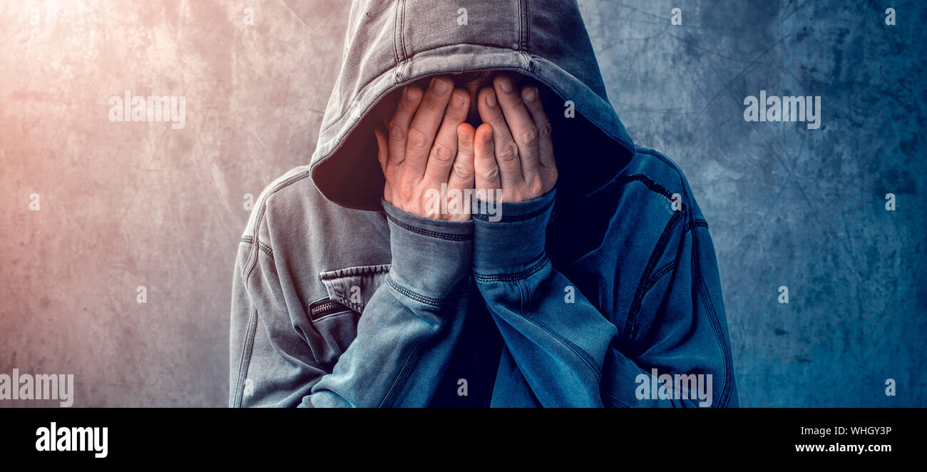 Desperate man in hooded jacket is crying, hands are covering face and tears in the eyes, light of hope shining from his right side Stock Photo