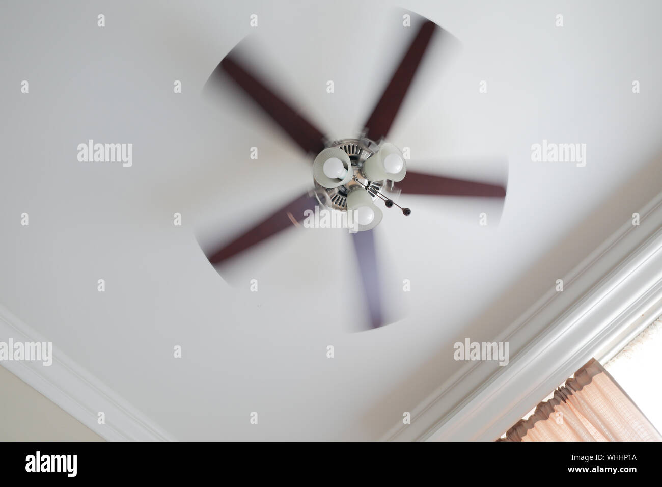 Ceiling fan with a ceiling lamp turned on, working with blurred blades in a living room, ceiling with moldings, curtain with shades and rods. Stock Photo