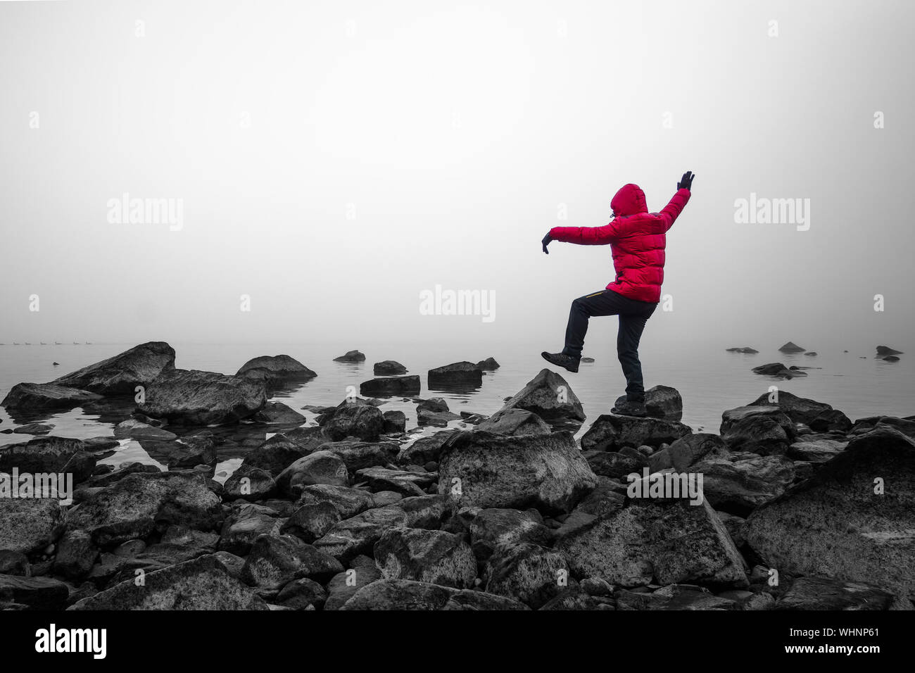 Rear View Full Length Of Playful Man In Parka On Rocks At Lakeshore During Foggy Weather Stock Photo