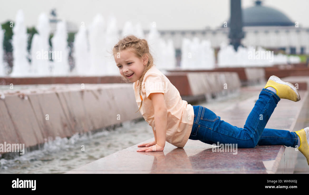 Cute little mischievous girl plays at the fountain. Time of prank. Urban casual outfit. Carefree. Happiness, fun and childhood concept. Stock Photo