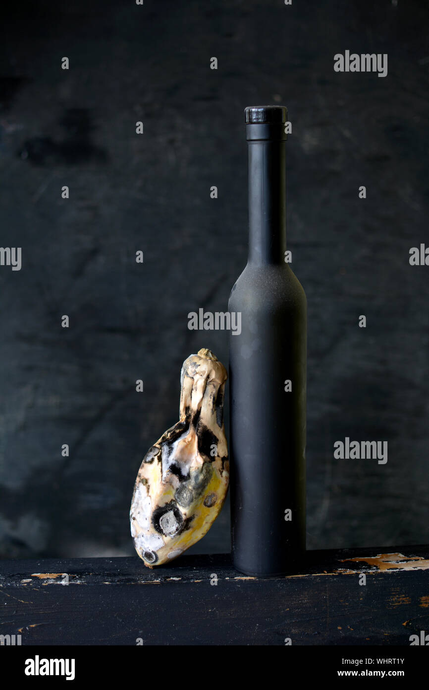 Still life with black wine bottle and rotten zucchini on dark background Stock Photo