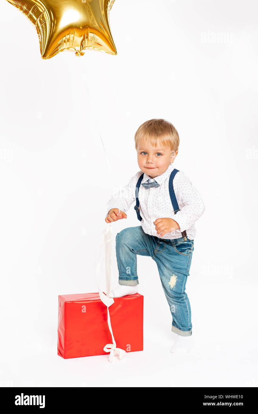Cute little boy, jeans and shirt, standing near present and blowing in the birthday tune. Gold balloon,shape star.Isolated on the white background Stock Photo