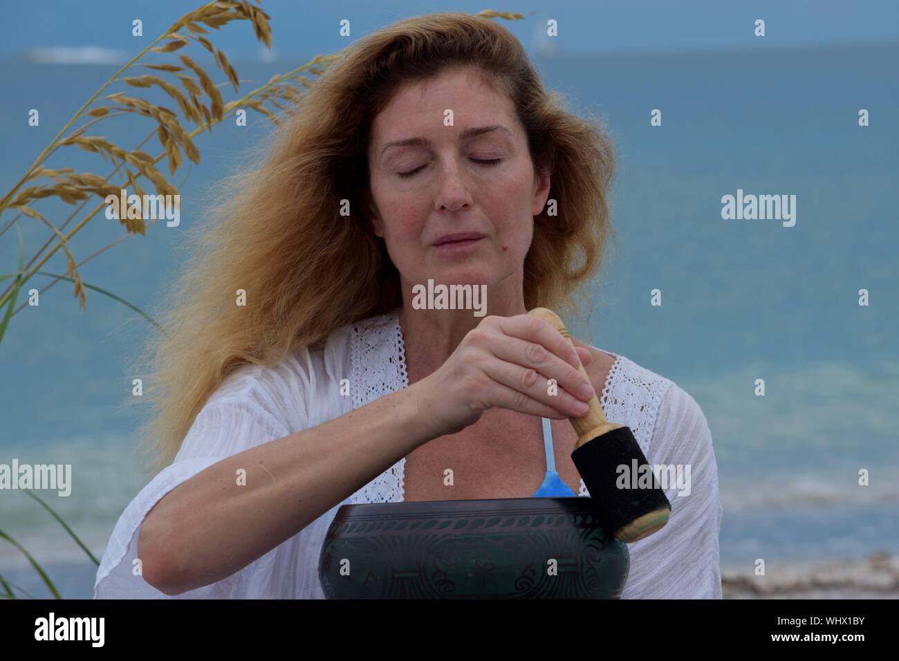 Woman Meditating With Singing Bowl With Wooden Striker At Beach Stock Photo