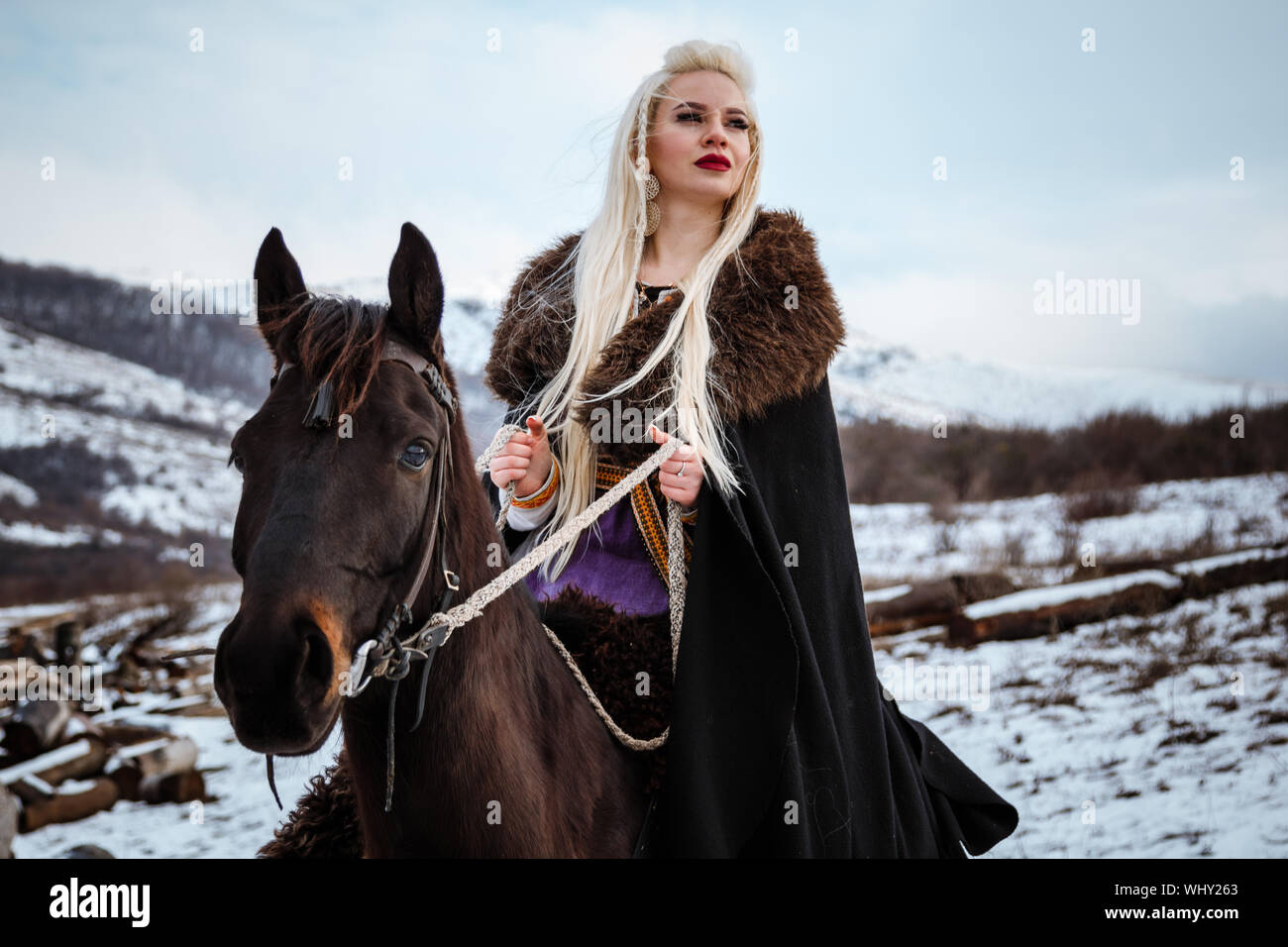 Beautiful young blonde on a crow. Woman viking with a black horse against the background of mountains Stock Photo