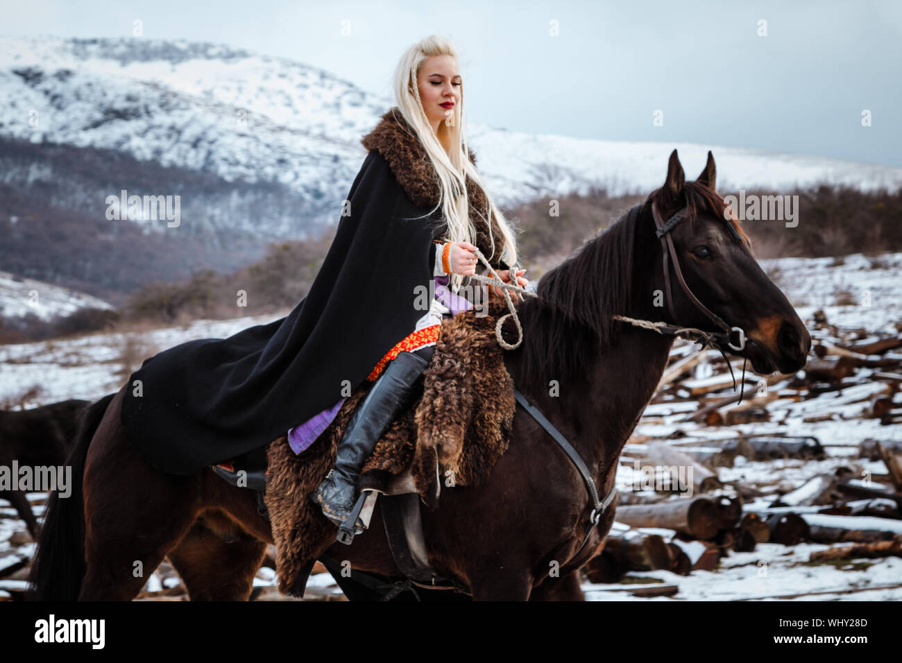 Beautiful young blonde on a crow. Woman viking with a black horse against the background of mountains Stock Photo