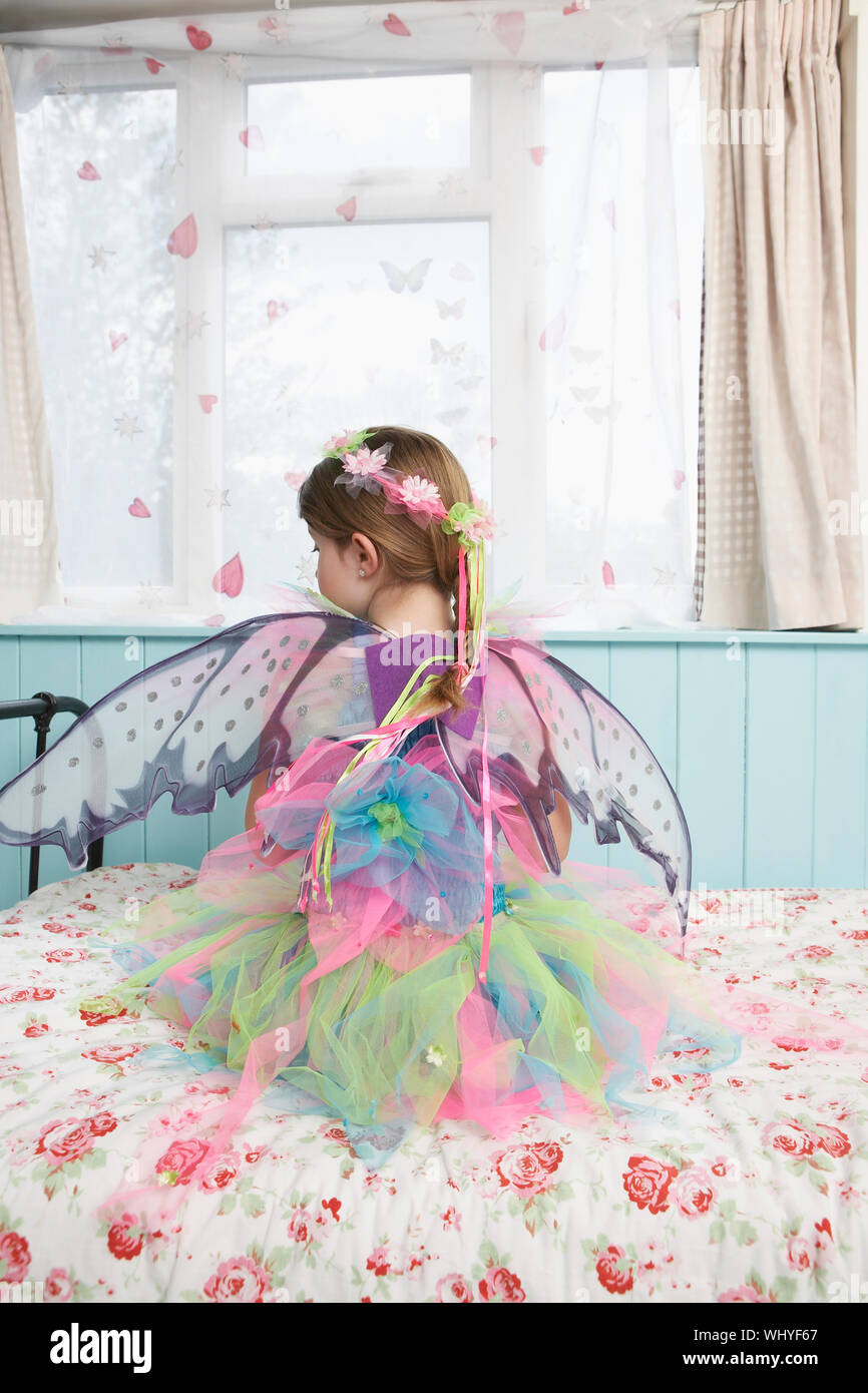 Rear view of a young girl in fairy costume sitting on bed looking ...