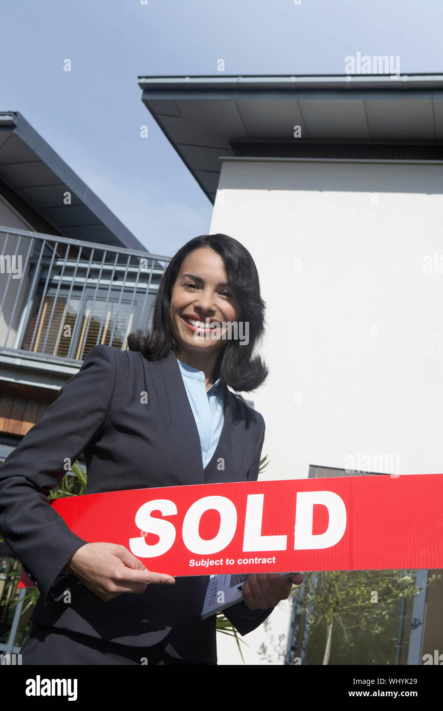 Portrait of a smiling female real estate agent holding sold sign ...