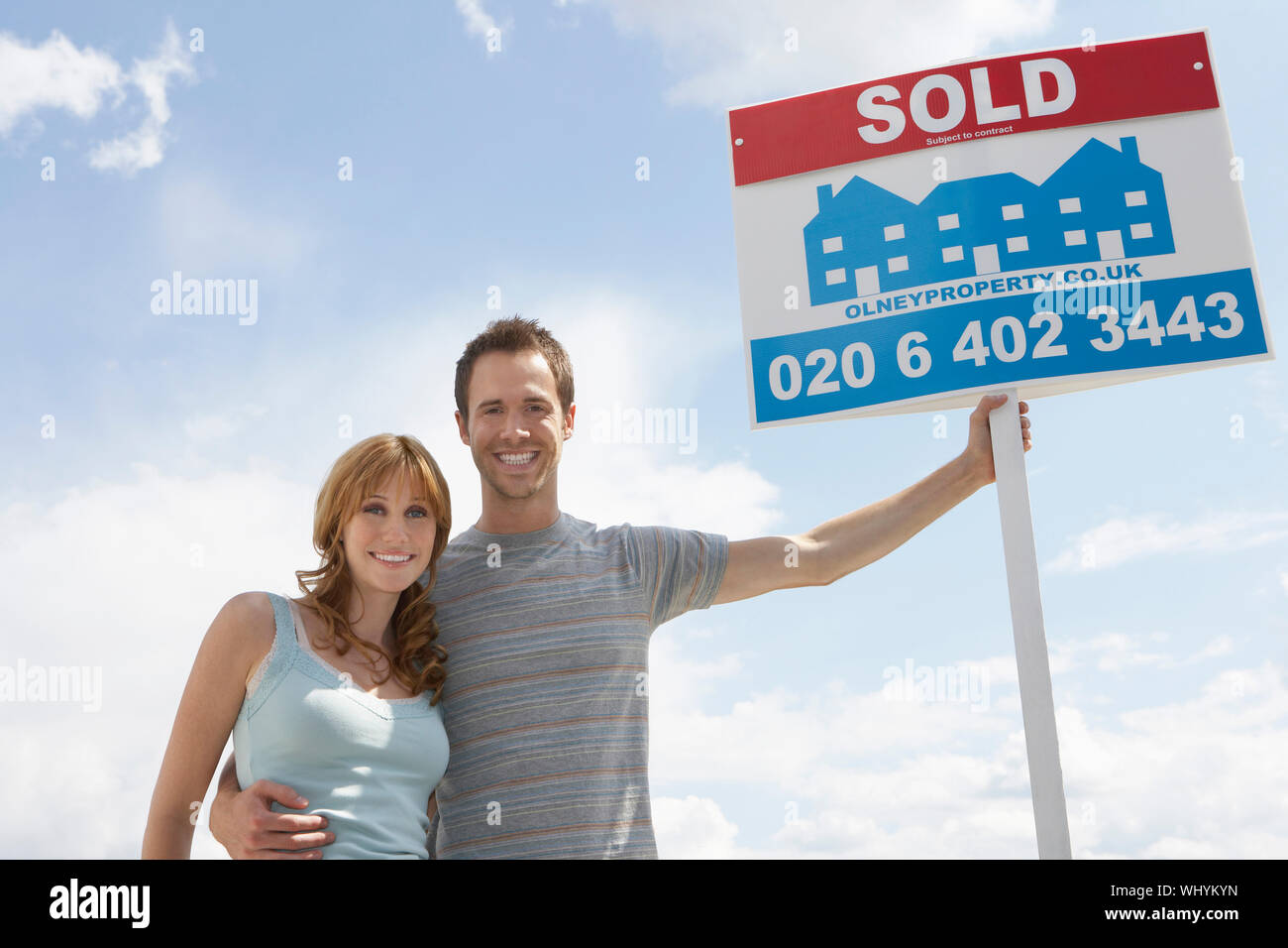 Portrait of a smiling young couple by sold sign standing against sky ...