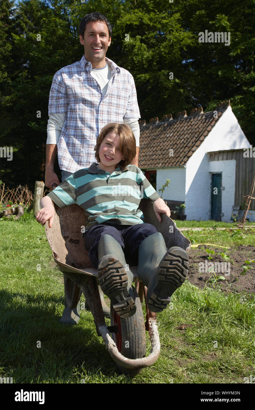 Portrait of a smiling man pushing son in wheelbarrow outside cottage Stock Photo