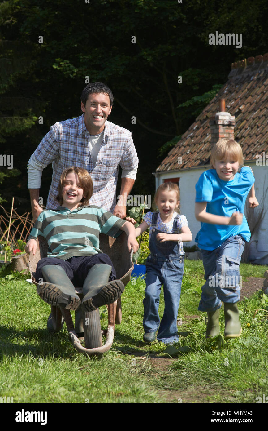 Full length of cheerful father and children racing with wheelbarrow outside cottage Stock Photo