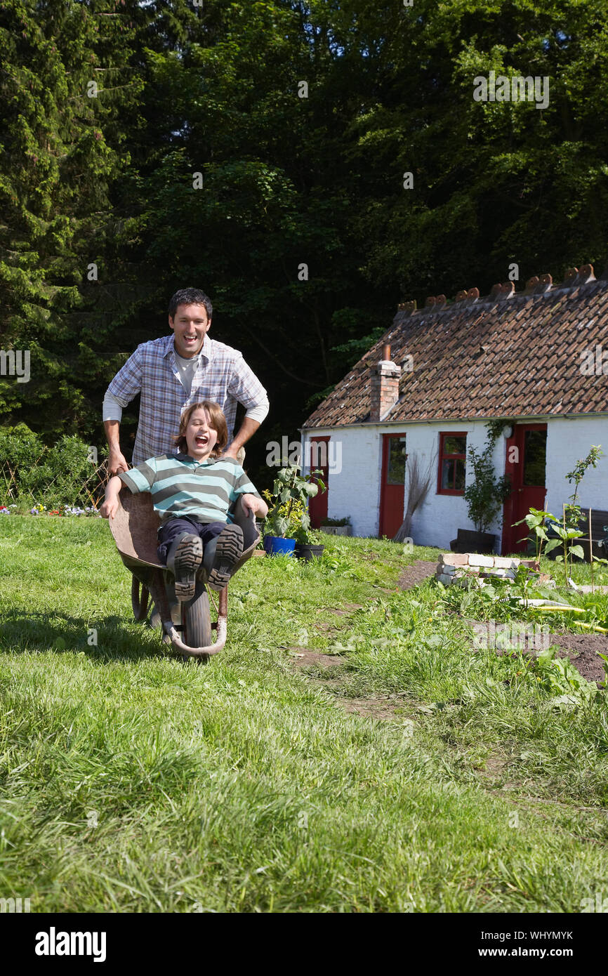 Portrait of a smiling man pushing son in wheelbarrow outside cottage Stock Photo