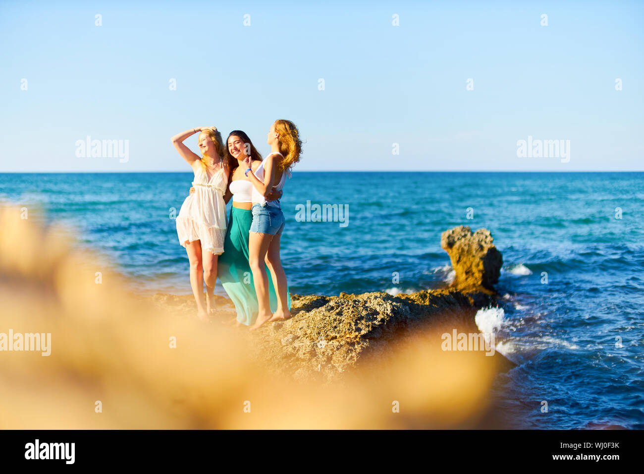 Happy Female Friends Standing On Rock By Sea During Sunny Day Stock Photo