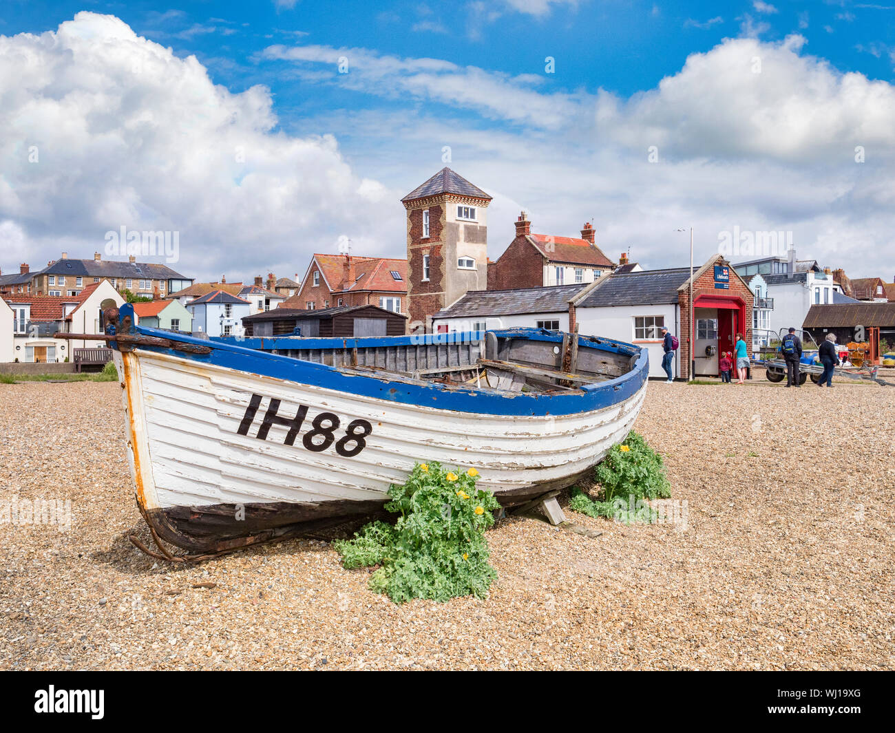16 June 2019: Aldeburgh, Suffolk, UK - Old boat on the beach, with the RNLI Lifeboat Station and people sightseeing. Sunny summer day. Stock Photo
