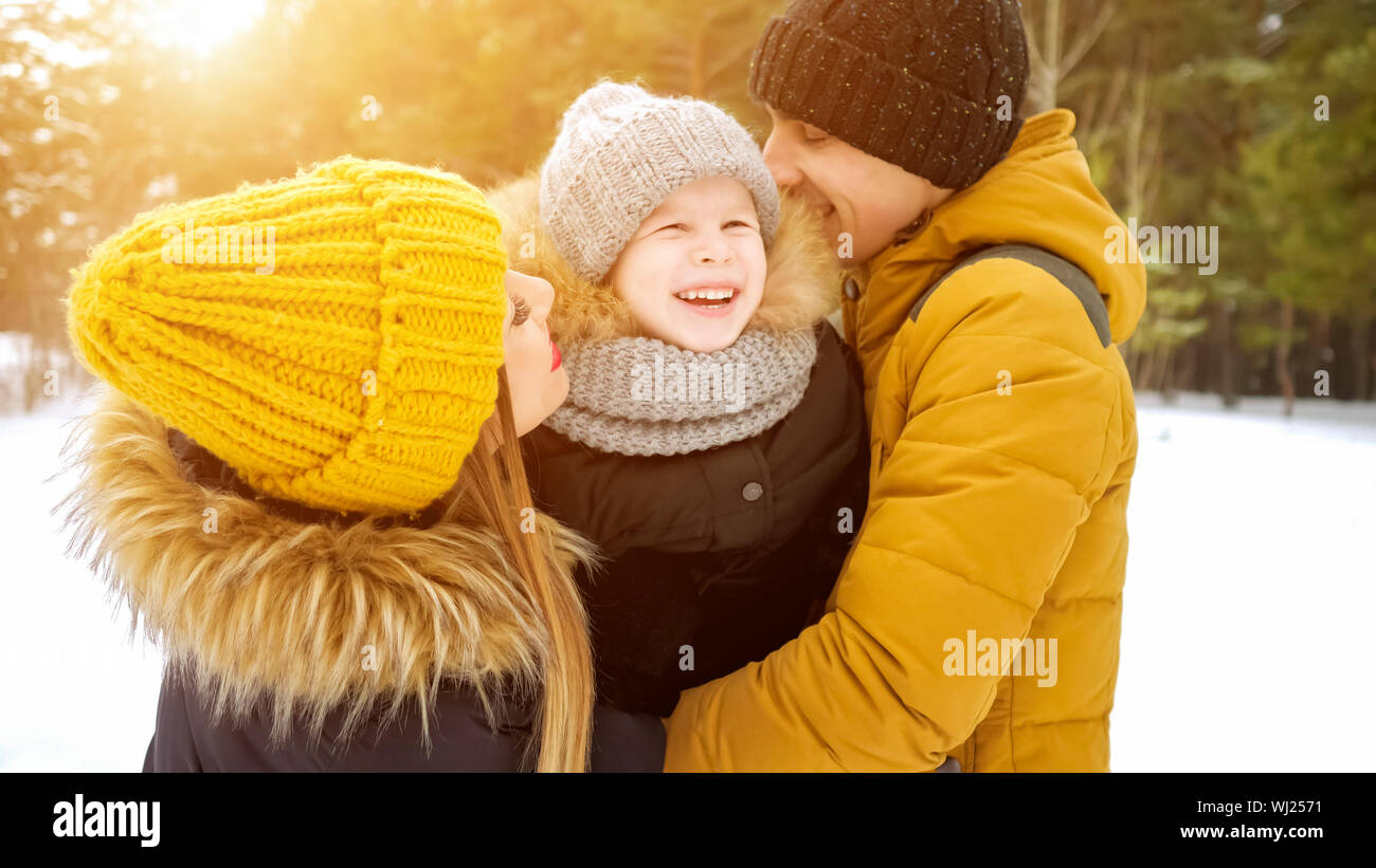 Portrait of happy family in winter day. Mom and dad are cuddling and kissing their little son in winter park. Family lovely moments. Stock Photo