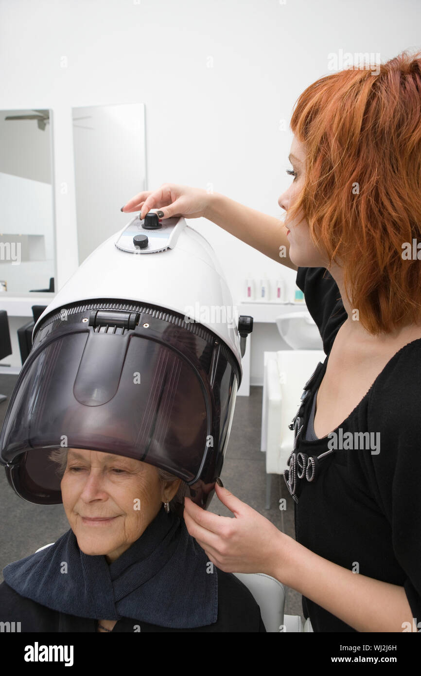 Senior woman under hooded hair dryer while stylist adjusting dial of it Stock Photo