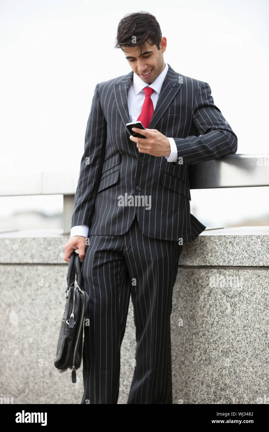 Young Indian businessman texting through cell phone while holding laptop bag at parapet Stock Photo