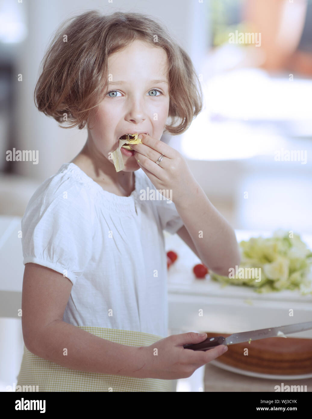 Young girl eating salad whilst holding knife in kitchen Stock Photo