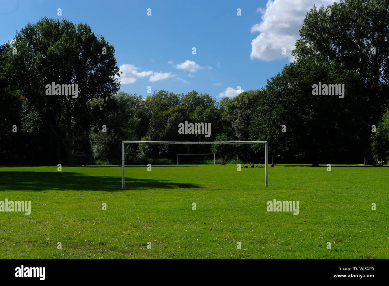 Two football or soccer goalposts in parallel in a park with green grass and blue skies and people sitting in the grass Stock Photo