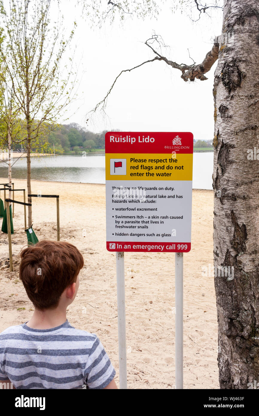 A young boy reading a warning sign to not enter the water at Ruislip Lido, Greater London, UK Stock Photo