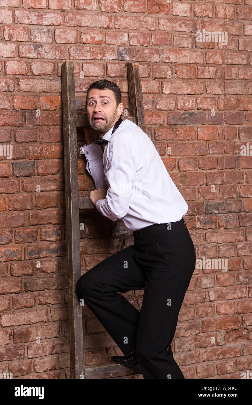 Terrified man trapped at the top of a ladder cowering against the brick wall with an expression or dread and fear Stock Photo