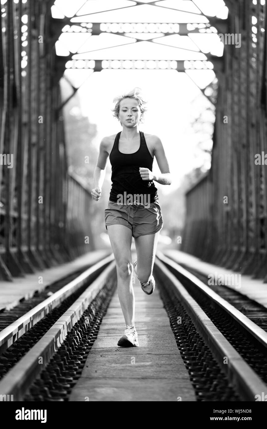 Athlete running on railaway tracks bridge in morning sunrise training for marathon and fitness. Healthy sporty caucasian woman exercising in urban env Stock Photo