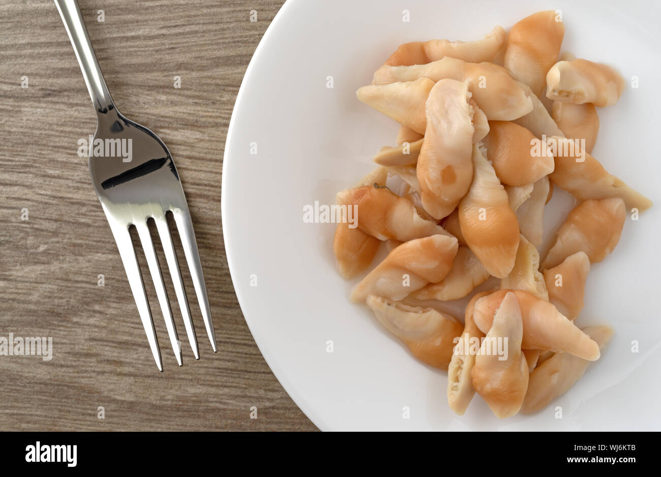 Top view of a serving of razor clams on a white plate with a fork to the side on a table illuminated with natural lighting. Stock Photo
