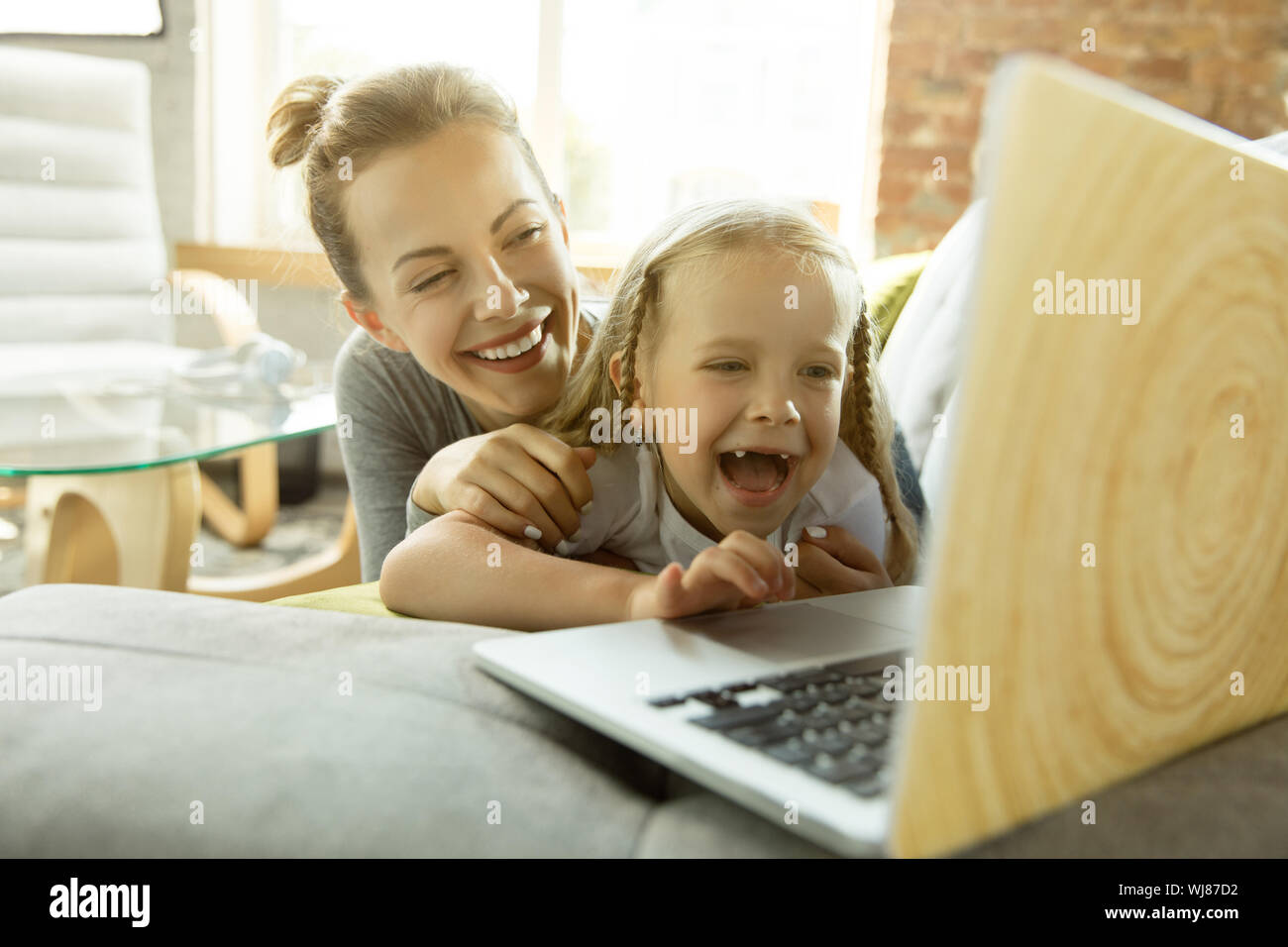 Female caucasian teacher and little girl, or mom and daughter. Homeschooling. Lying on the sofa and using laptop for knowledges getting while lesson is. Education, school, studying concept. Stock Photo