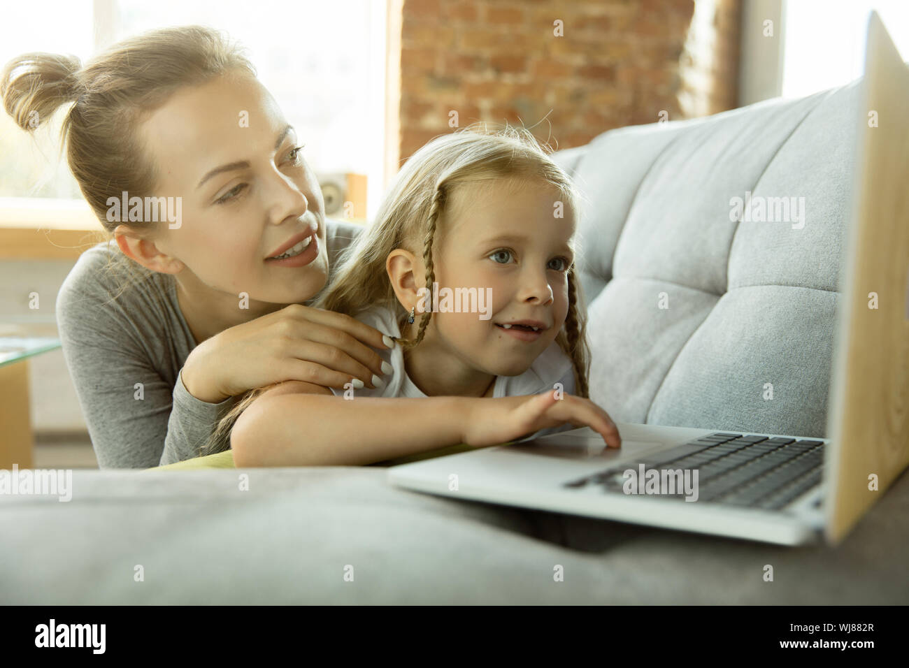 Female caucasian teacher and little girl, or mom and daughter. Homeschooling. Lying on the sofa and using laptop for knowledges getting while lesson is. Education, school, studying concept. Stock Photo