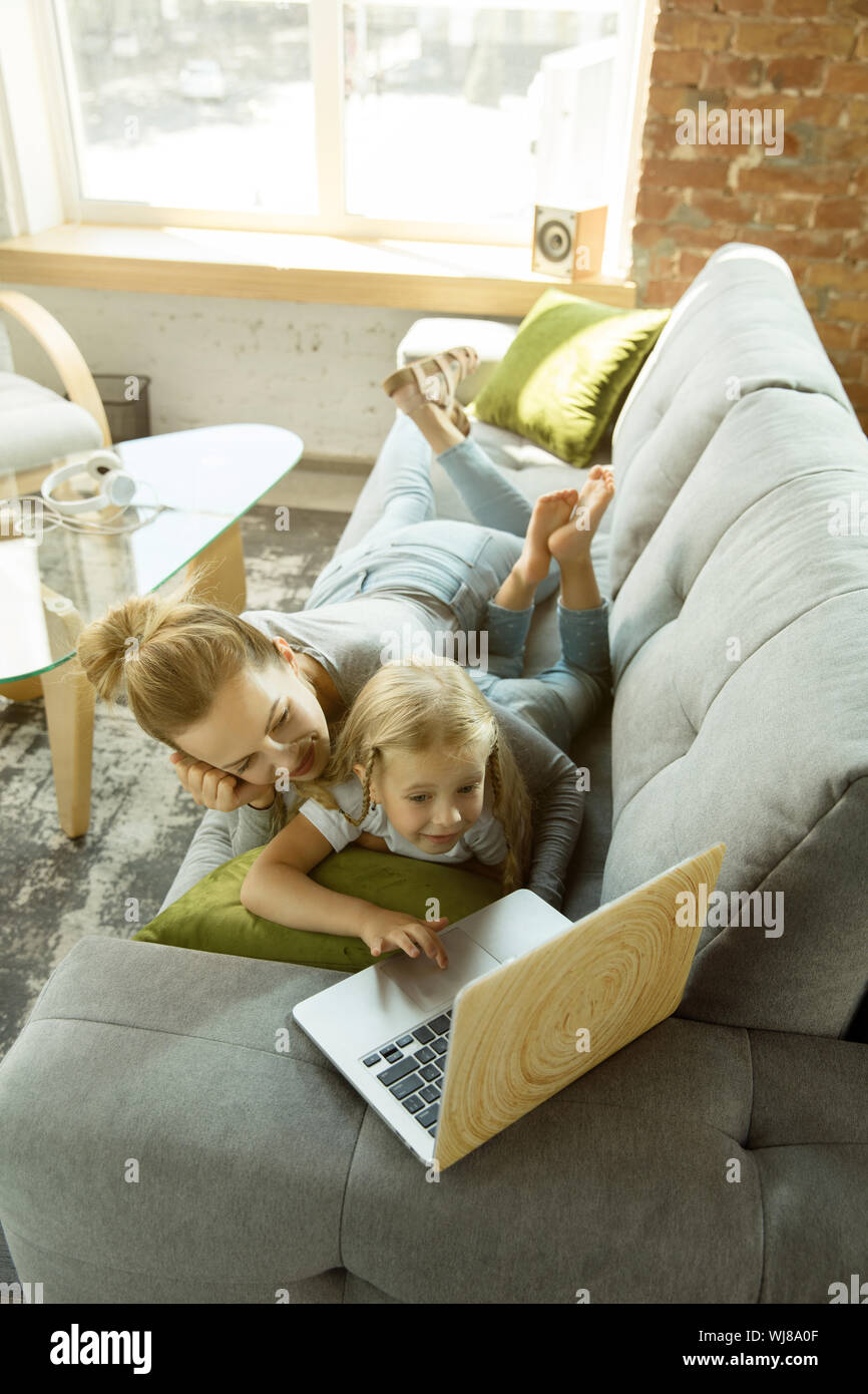 Female caucasian teacher and little girl, or mom and daughter. Homeschooling. Lying on the sofa and using laptop for knowledges getting while lesson is. Education, school, studying concept. Stock Photo