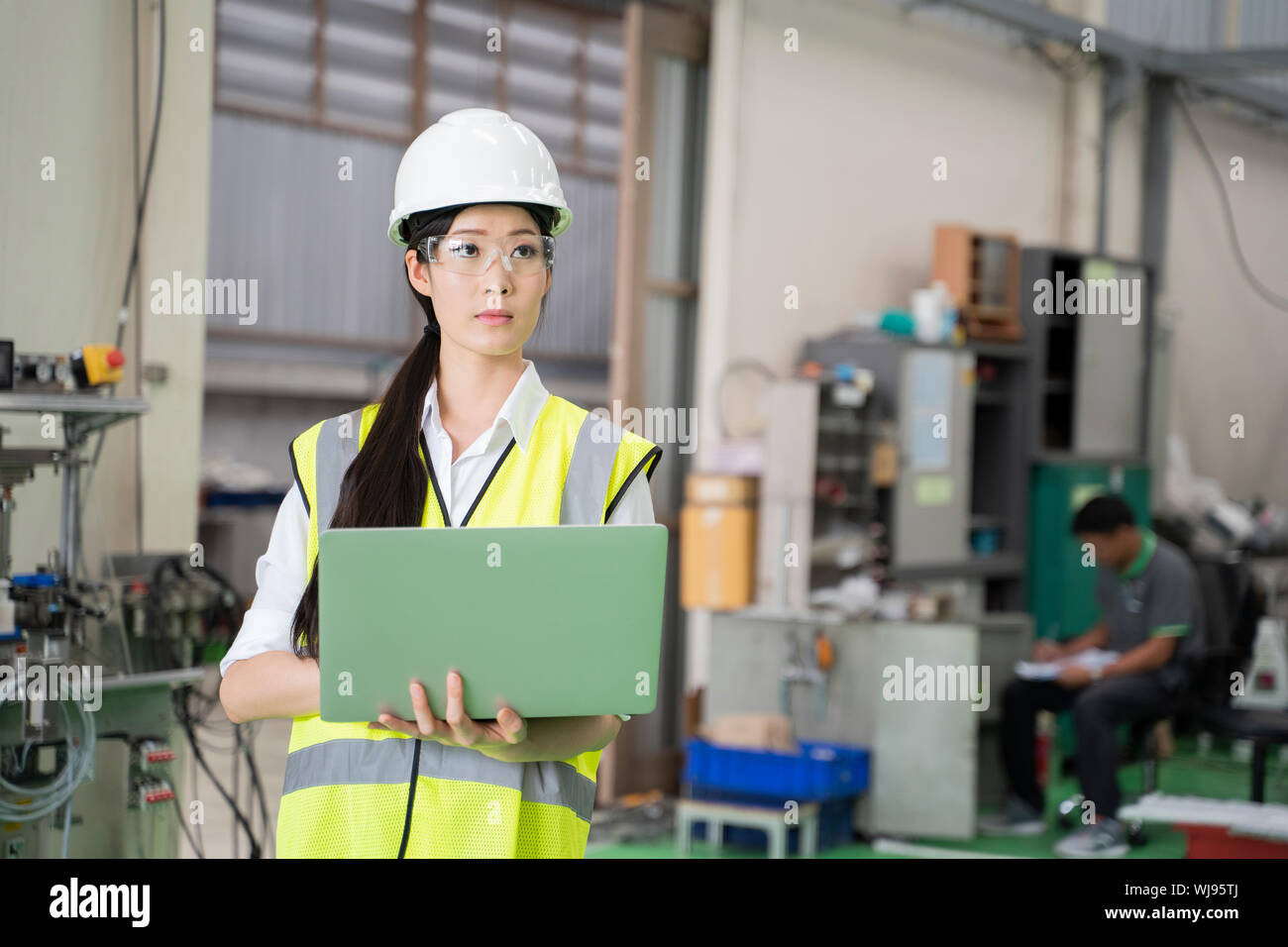 Female Industrial Engineer in the Hard Hat Uses Laptop Computer while Standing in the Industry Factory. In the Background Various Metal work Project P Stock Photo