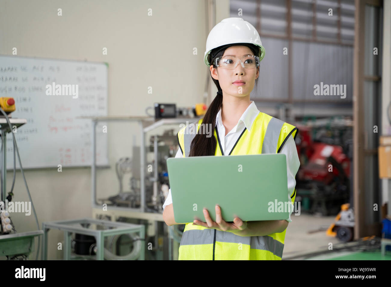 Female Industrial Engineer in the Hard Hat Uses Laptop Computer while Standing in the Industry Factory. In the Background Various Metal work Project P Stock Photo