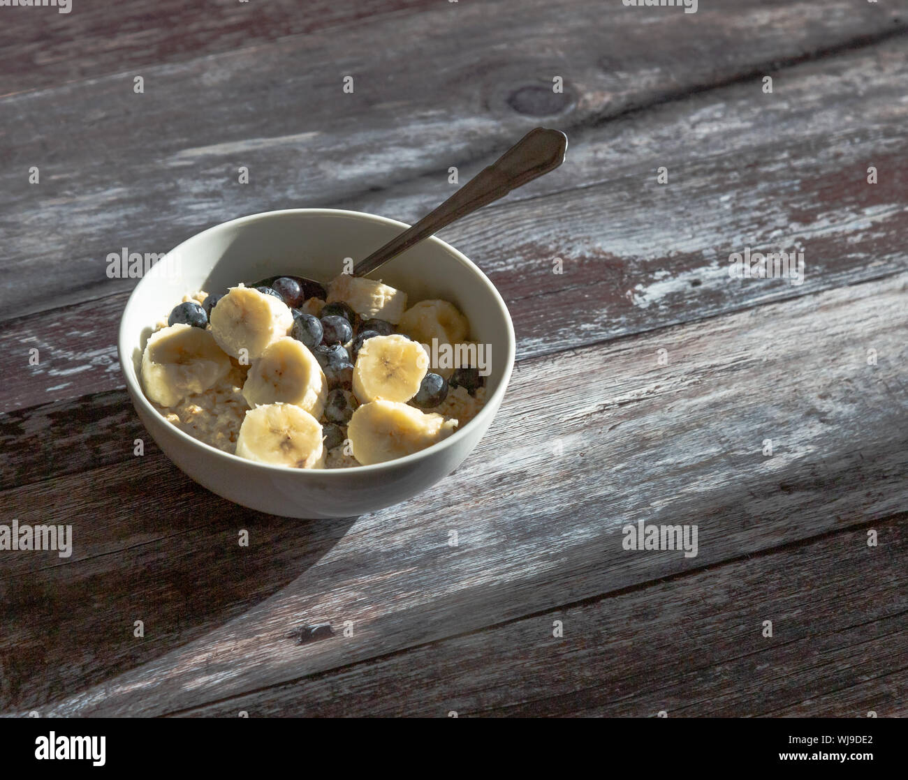 A bowl of oat porridge topped with sliced banana and blueberries. The bowl is on a rustic wooden background allowing plenty of copy space. Stock Photo