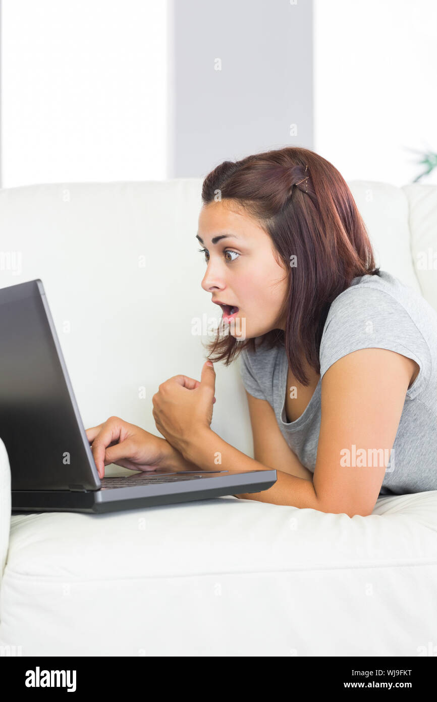 Brunette young woman using her notebook lying taken aback in front of it at home in the living room Stock Photo