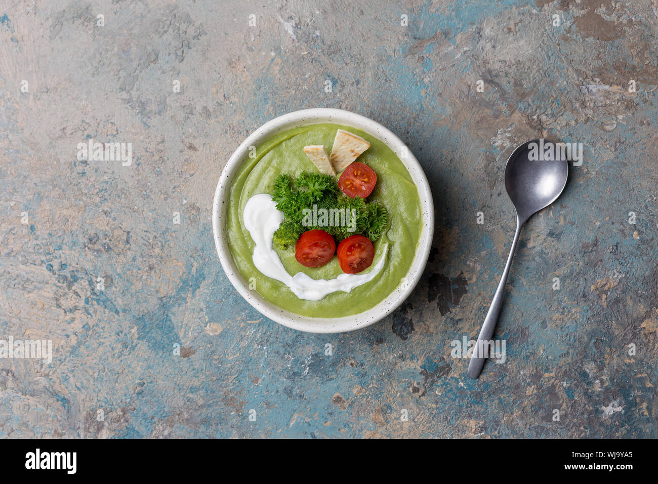 Bowl with broccoli cream soup, parsley, cherry tomatoes, pita and spoon on blue and green concrete background. Top view, table top, flat lay Stock Photo