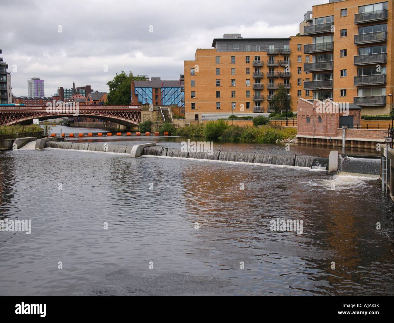 Red bridge Orange boom weir side flow on the river Aire in Leeds with modern flats Leeds Yorkshire England Stock Photo