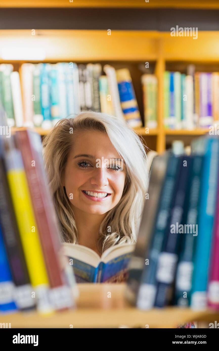 Portrait of a smiling young female student reading book amid bookshelves in the college library