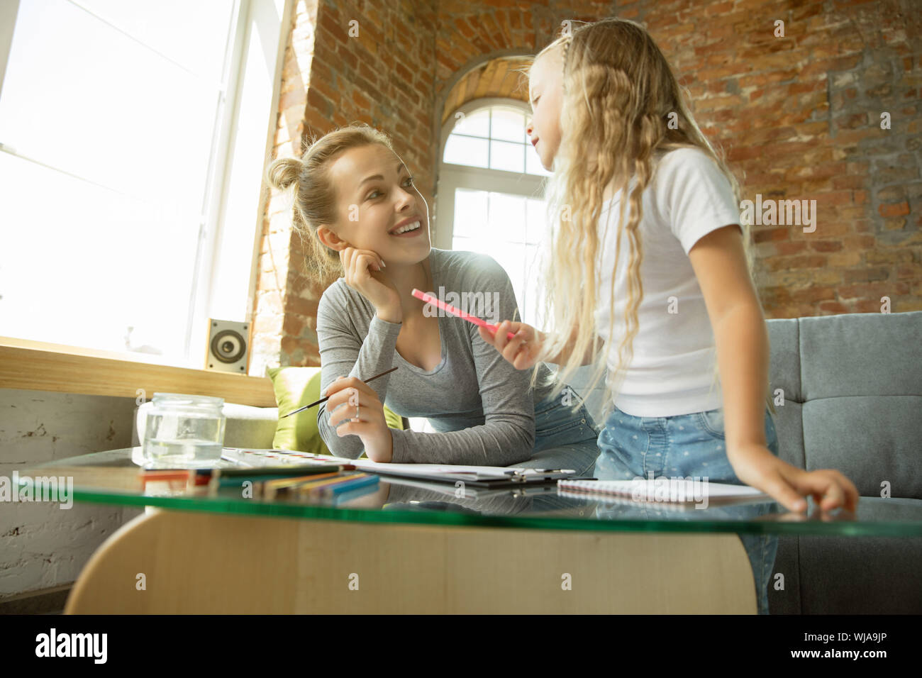 Female caucasian teacher and little girl, or mom and daughter. Homeschooling. Sitting on the sofa and drawing with paints, writing, talking and having fun. Education, school, studying concept. Stock Photo