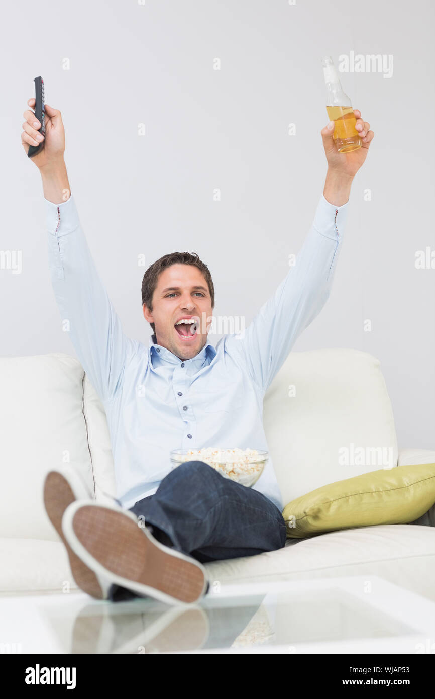 Man with a drink and remote control cheering on sofa Stock Photo
