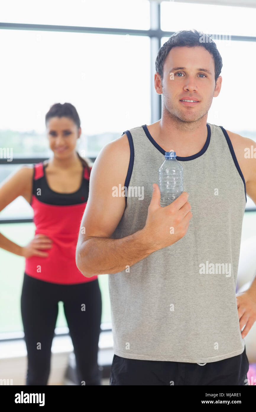 Fit man holding water bottle with friend in background in exercise room Stock Photo