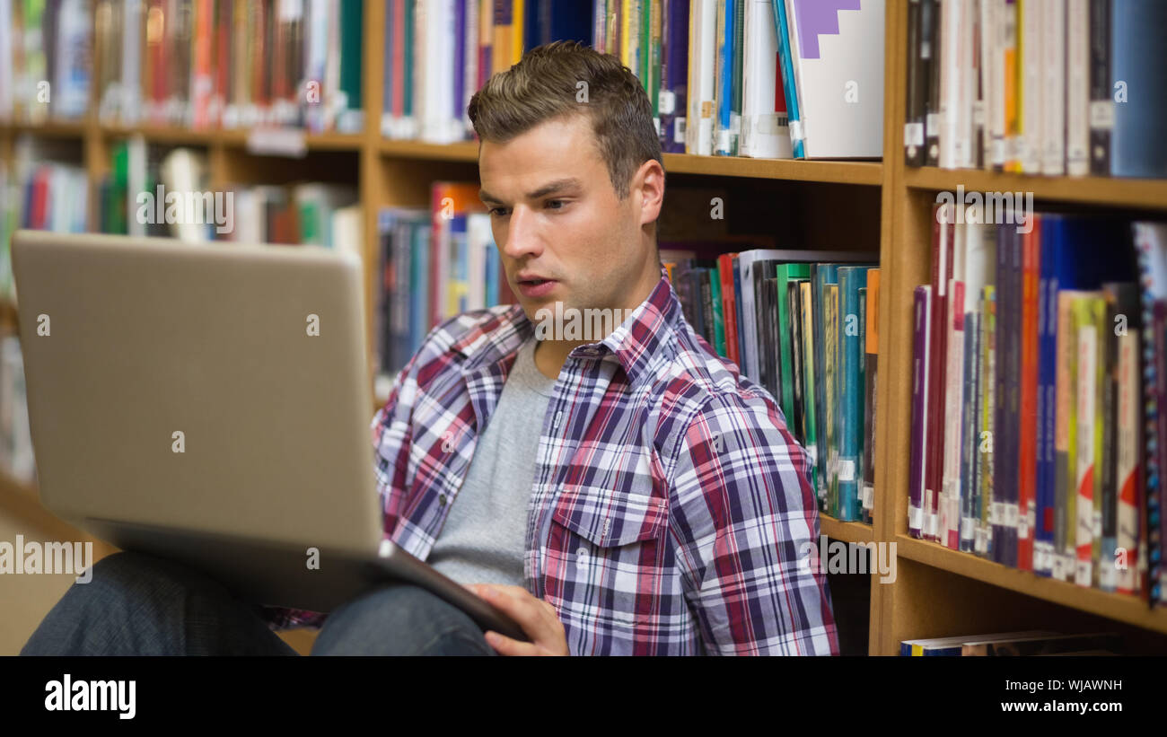 Serious young student sitting on library floor using laptop Stock Photo