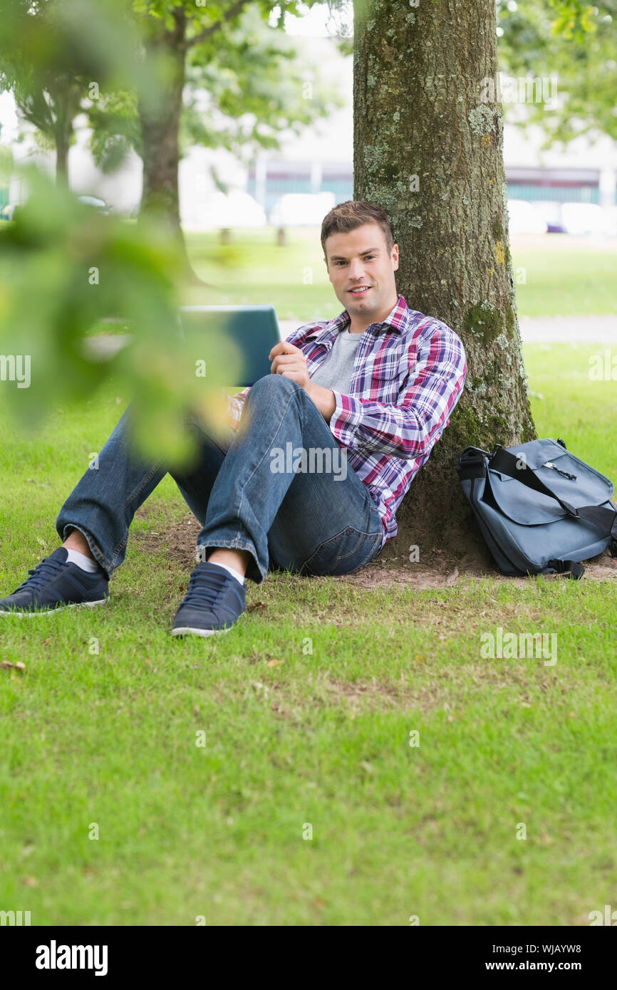 Smiling student using his tablet pc outside leaning on tree Stock Photo
