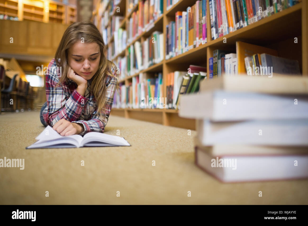 Pretty focused student lying on library floor reading book Stock Photo