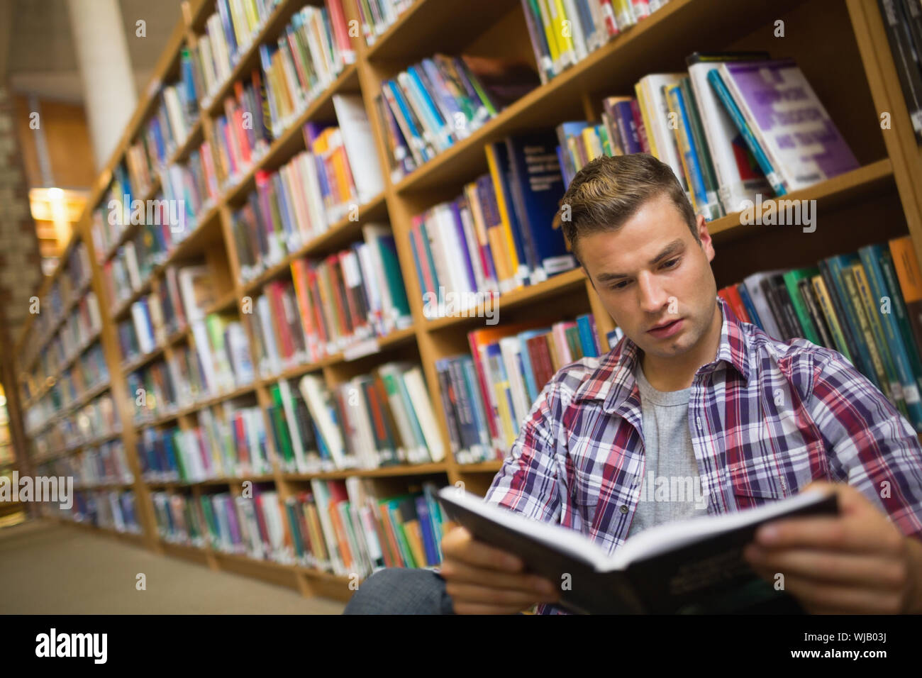 Focused young student sitting on library floor reading book Stock Photo